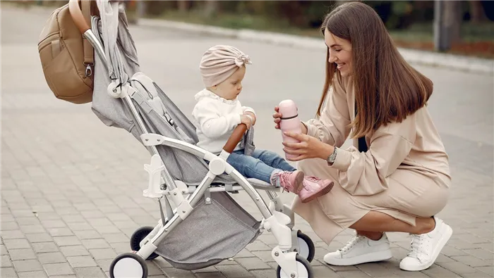 Маленькая девочка в белой коляске, а рядом мама с бутылочкой.